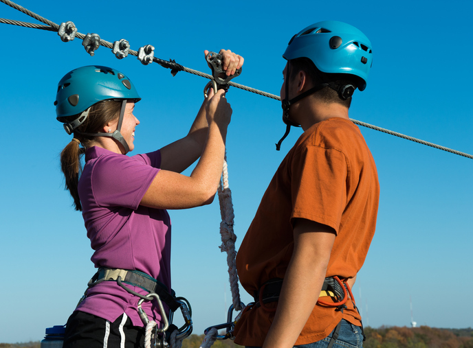 two adults getting ready to go on a zip line with equipment and helmets